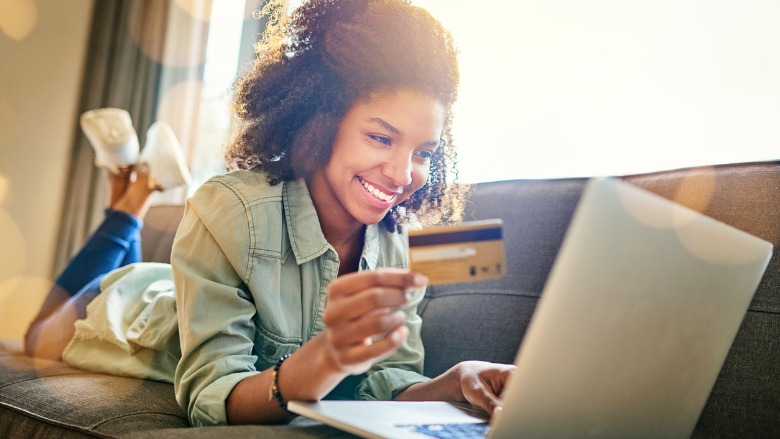 Woman laying on couch with credit card looking at laptop